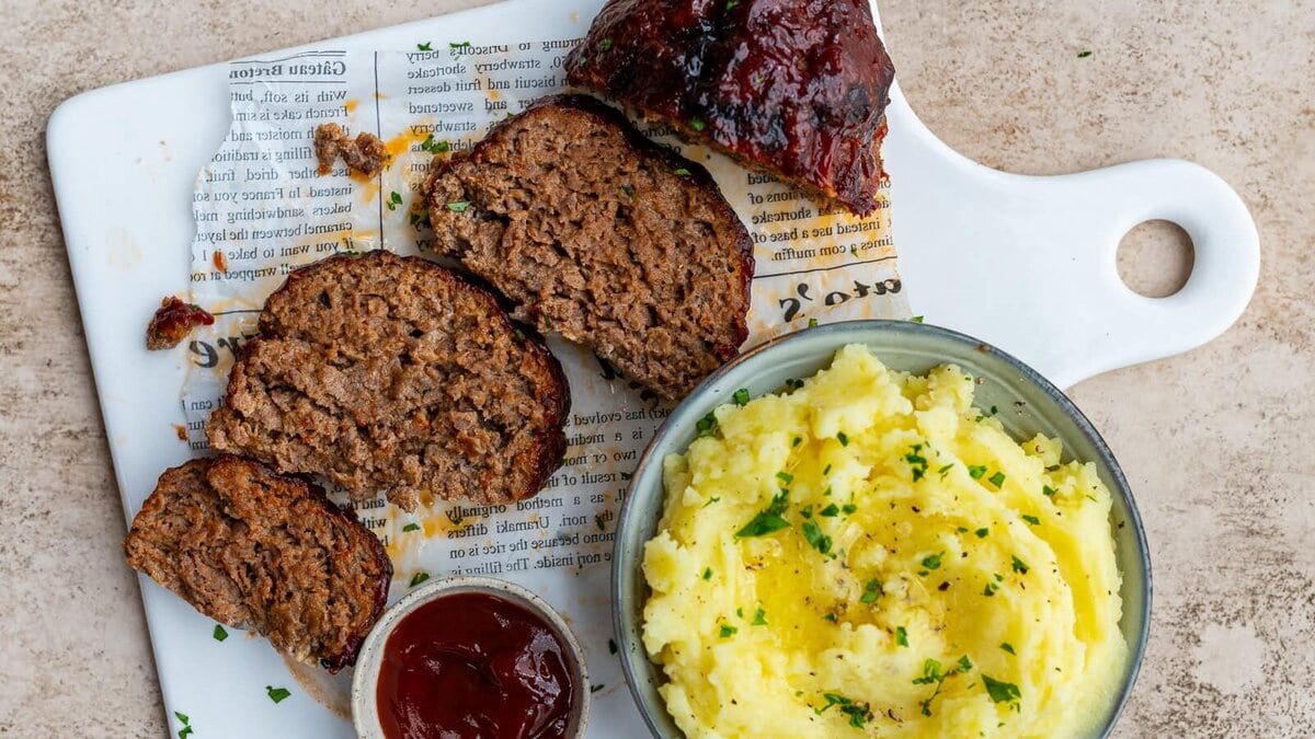 A white cutting board holds sliced meatloaf with glaze, mashed potatoes garnished with herbs in a bowl, and a small container of ketchup, all arranged on a piece of printed paper.
