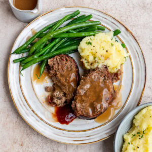 A plate with two slices of air fryer meatloaf topped with brown gravy, mashed potatoes with herbs, and green beans. Nearby are a small jug of gravy and a bowl of extra mashed potatoes.