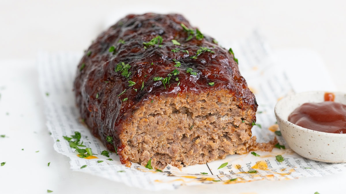 A sliced loaf of glazed meatloaf garnished with chopped parsley sits on a piece of printed parchment, next to a small bowl of ketchup.