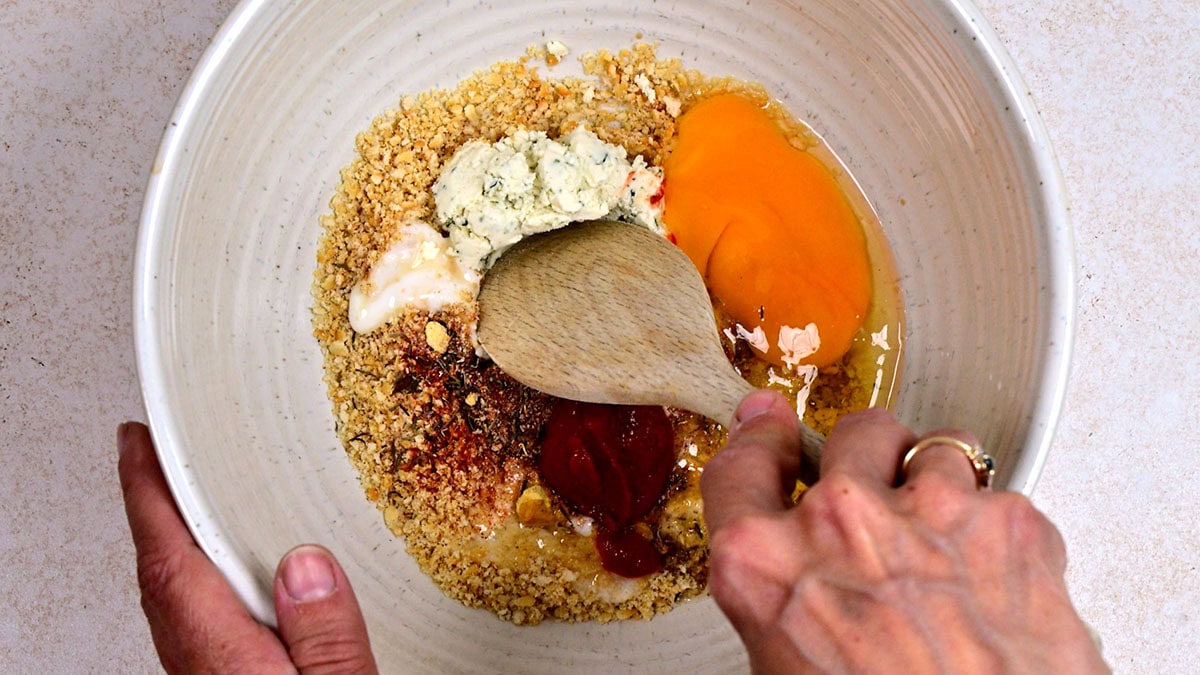 A hand holds a wooden spoon mixing ingredients including breadcrumbs, an egg, ketchup, spices, and a creamy mixture in a large white bowl on a light countertop.