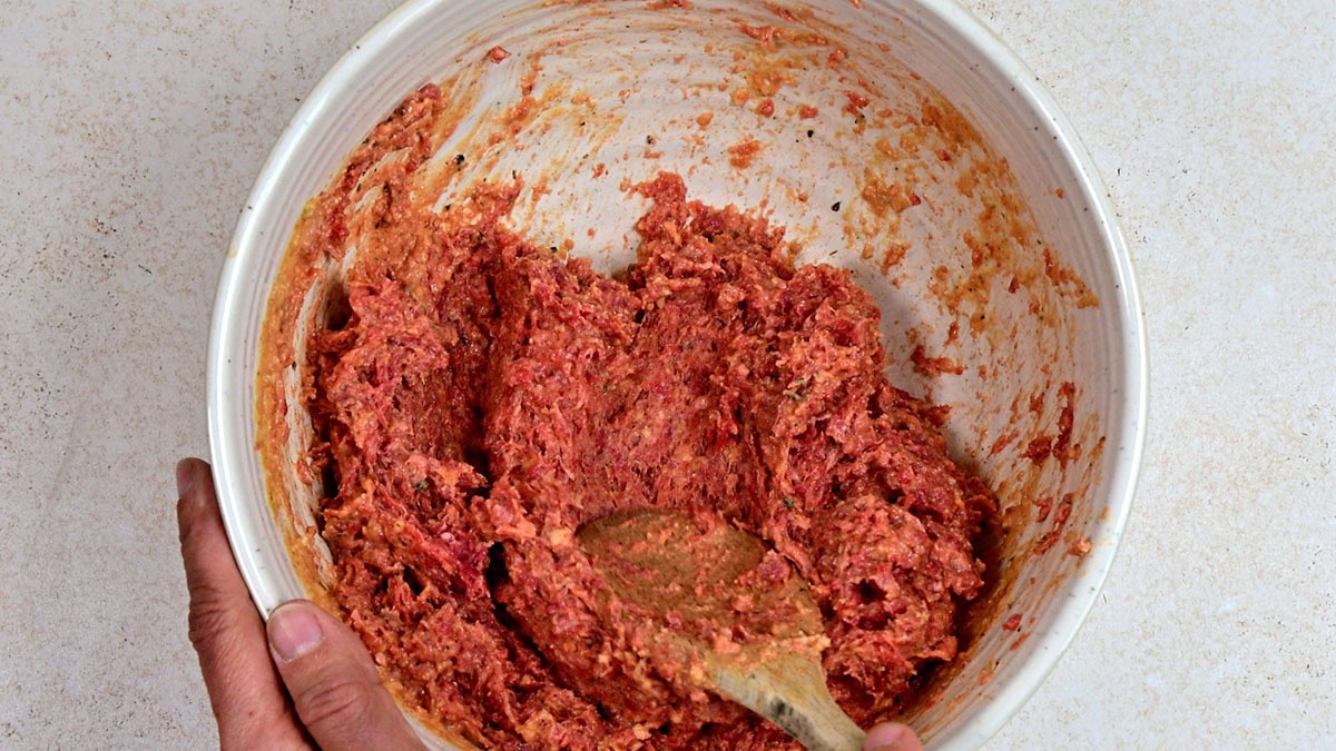A person is mixing raw ground meat with seasonings in a white bowl using a wooden spoon. The mixture appears to be for making meatballs or meatloaf.