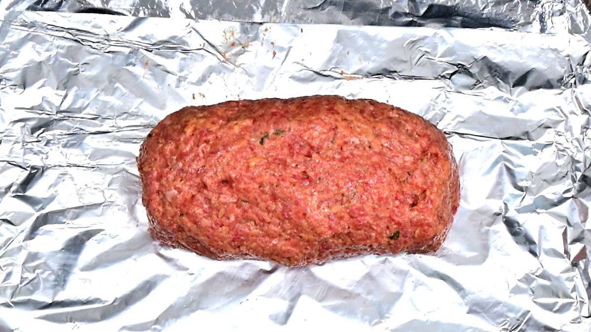 A raw, oval-shaped meatloaf mixture sits on a sheet of crinkled aluminum foil, ready to be baked.