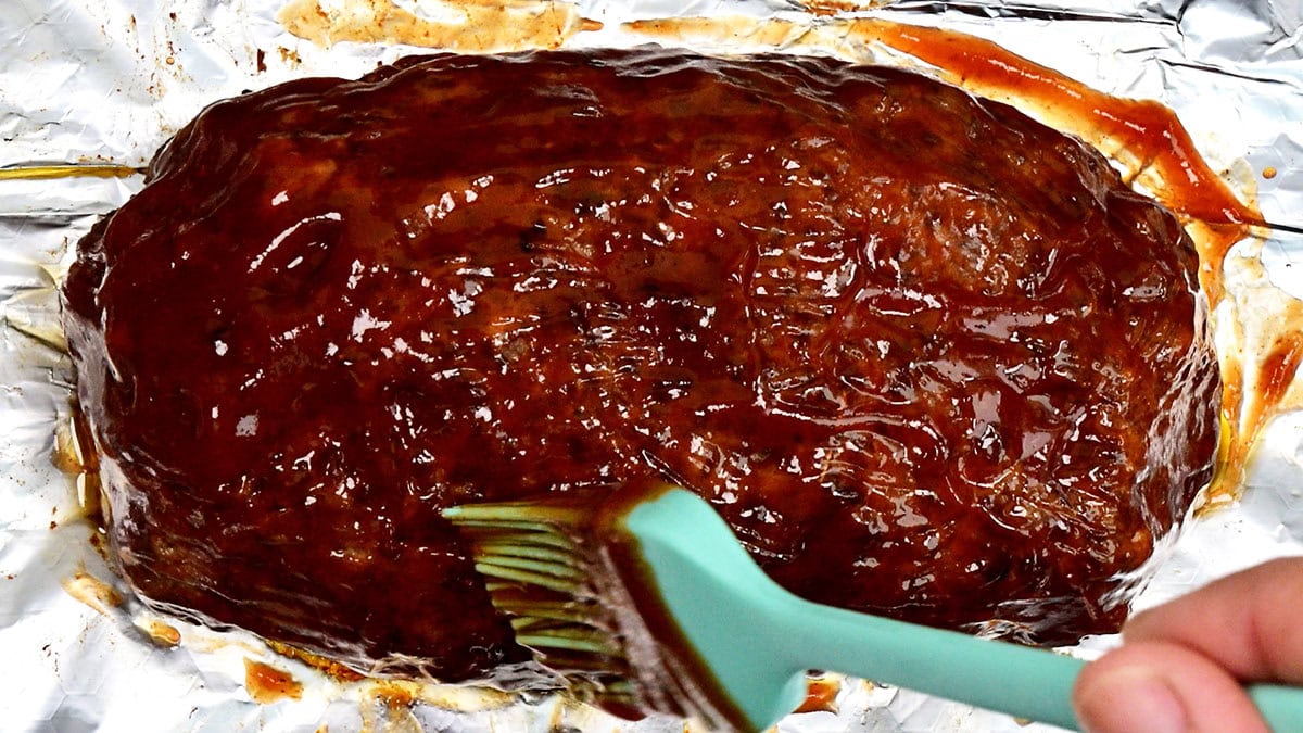 A hand uses a silicone brush to spread sauce over a cooked meatloaf resting on a sheet of aluminum foil.