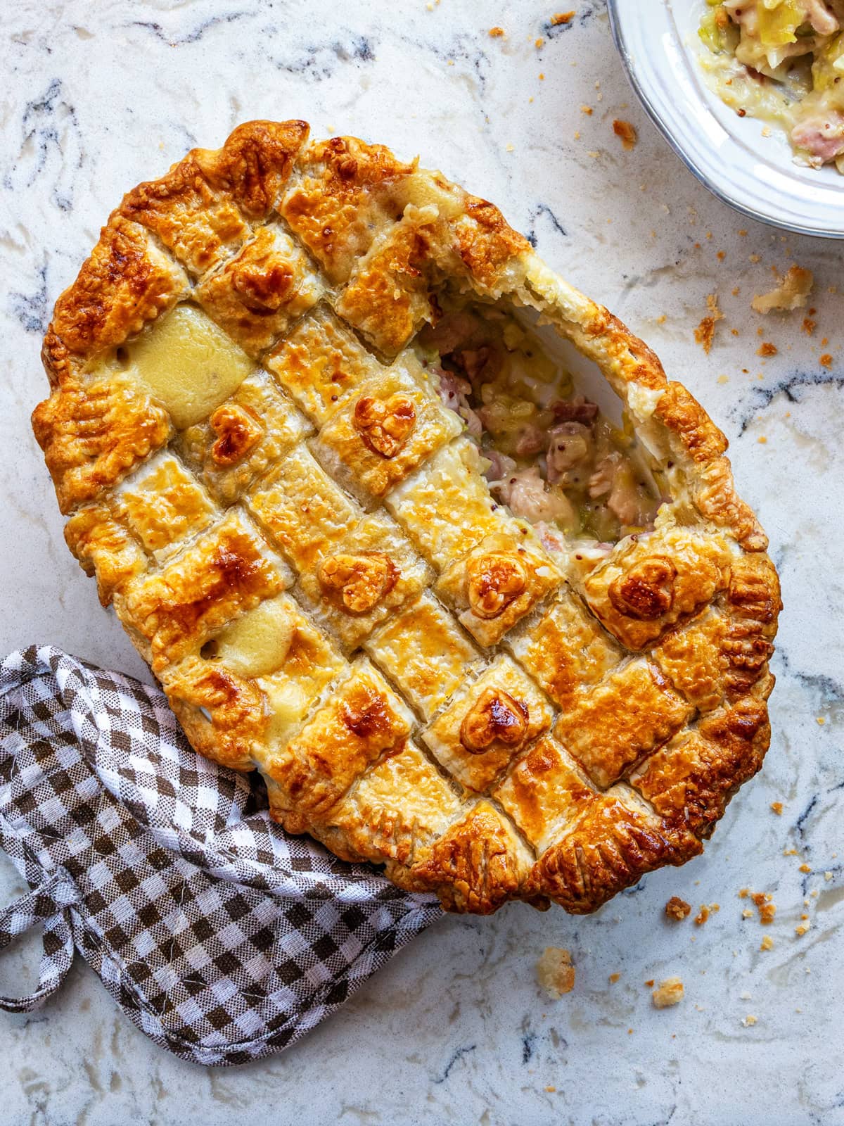 A golden-brown lattice chicken and leek pie with a portion missing, revealing a creamy filling of chicken and vegetables, sits on a marble surface next to a brown checkered cloth.