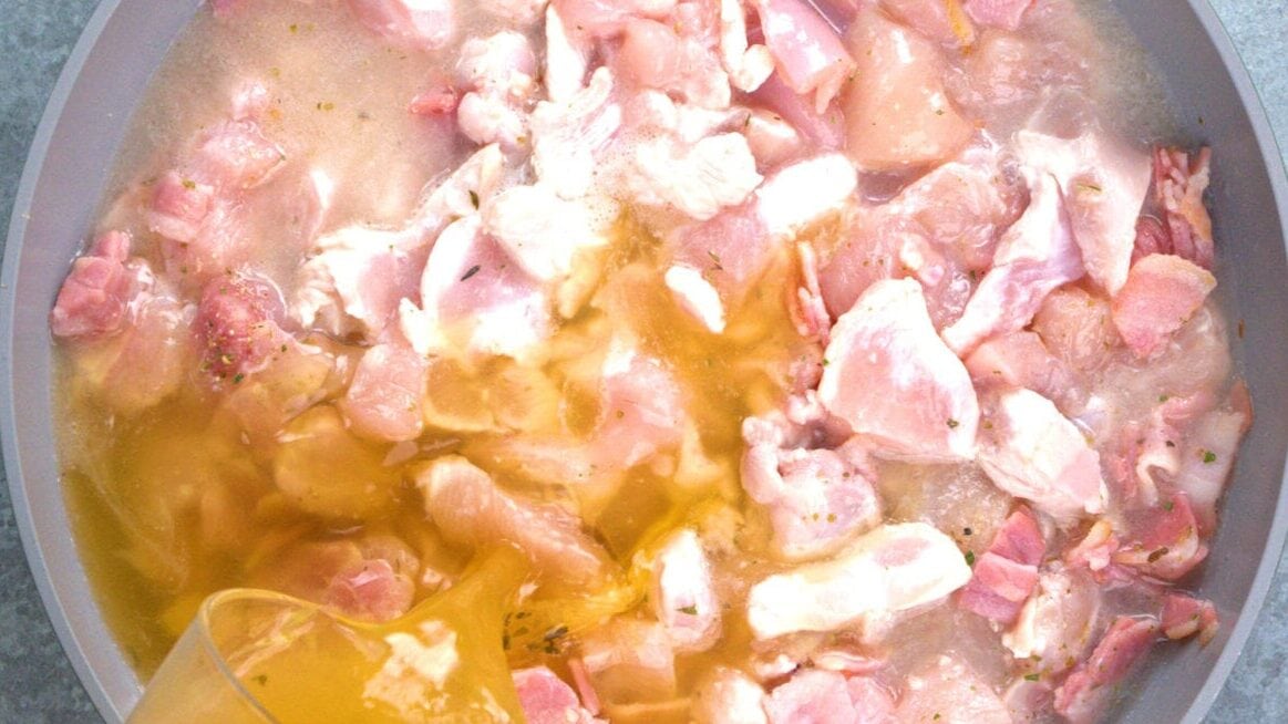 Raw, chopped chicken pieces in a pan with broth being poured over them, preparing for cooking.