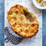Golden-brown chicken and leek pie with a lattice puff pastry topping with a slice removed, revealing creamy filling, sits on a marble board next to a checkered oven mitt and a bowl of filling on a blue-gray surface.