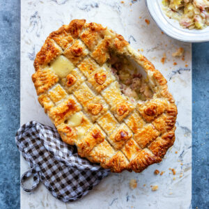 Golden-brown chicken and leek pie with a lattice puff pastry topping with a slice removed, revealing creamy filling, sits on a marble board next to a checkered oven mitt and a bowl of filling on a blue-gray surface.