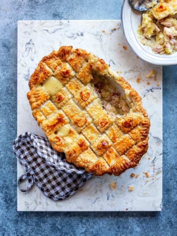 Golden-brown chicken and leek pie with a lattice puff pastry topping with a slice removed, revealing creamy filling, sits on a marble board next to a checkered oven mitt and a bowl of filling on a blue-gray surface.
