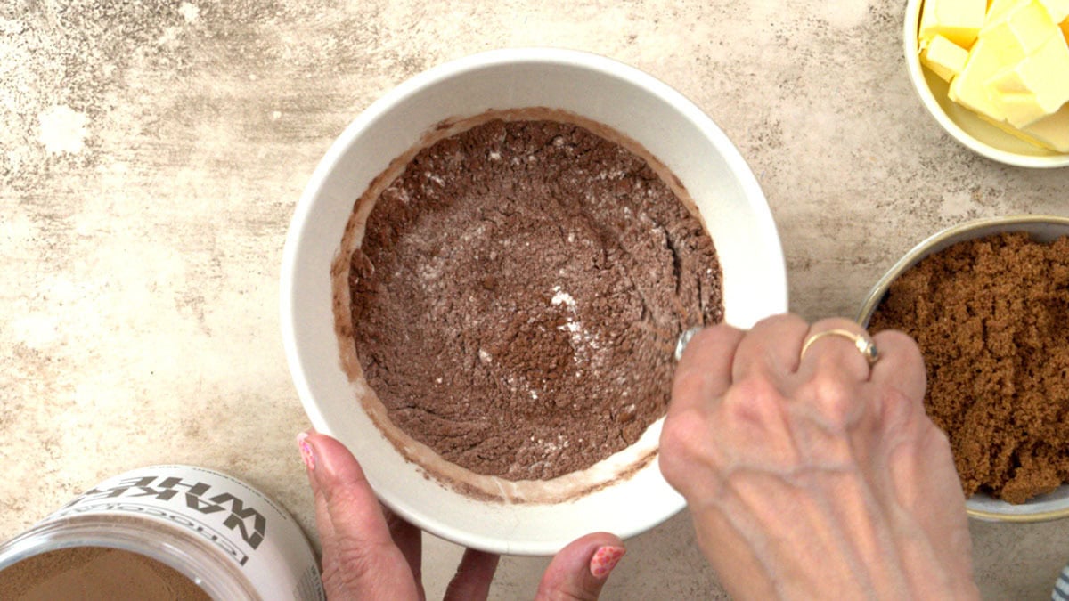 A person stirs a bowl of dry cocoa powder mixture on a countertop. Nearby are bowls containing brown sugar and butter, and a container labeled Naked Nut.