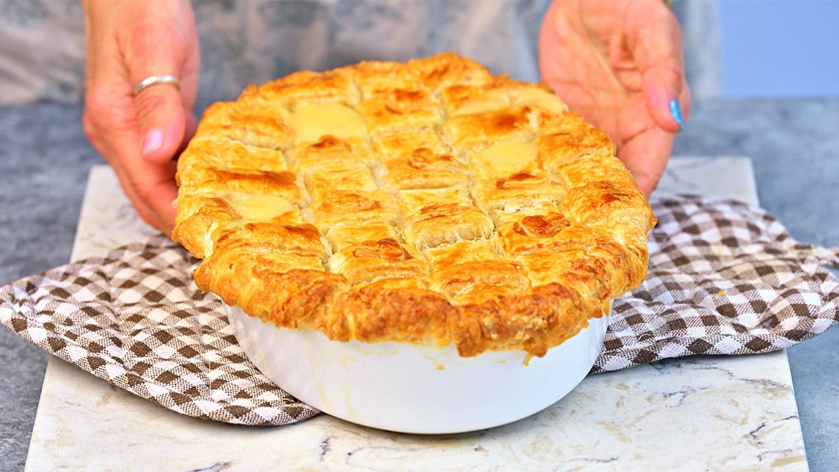 A person places a freshly baked pie with a golden, flaky lattice crust on a checkered cloth, the pie resting in a white ceramic dish on a marbled surface.