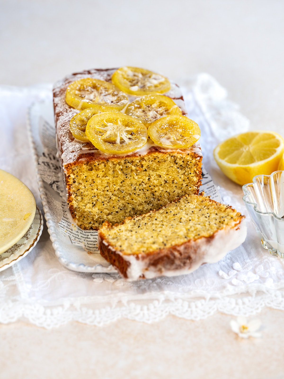 A loaf of lemon poppy seed cake topped with glazed lemon slices sits on a tray with a slice cut. A halved lemon, glass dish, and a small plate are nearby, all on a lace cloth.