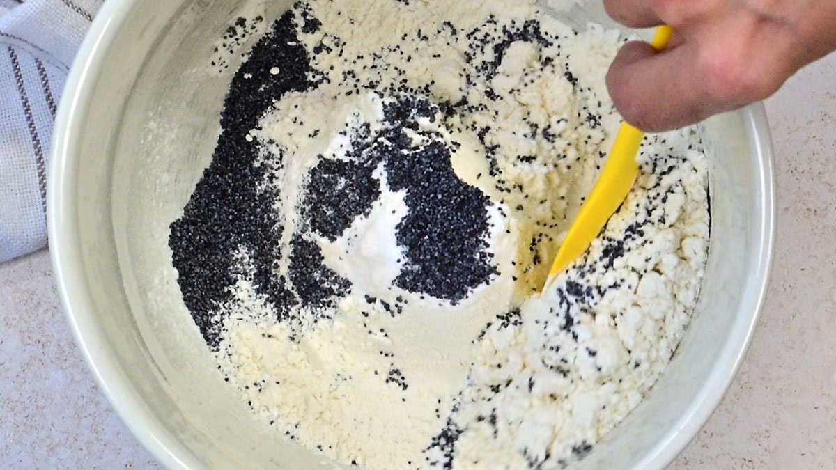A hand uses a yellow spatula to mix flour and poppy seeds in a white bowl, with a striped towel partly visible beside the bowl.