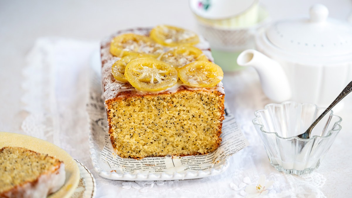 A loaf of lemon poppy seed bread topped with candied lemon slices sits on a tray lined with paper. In the background, there are teacups, a white teapot, and a glass bowl with a spoon.