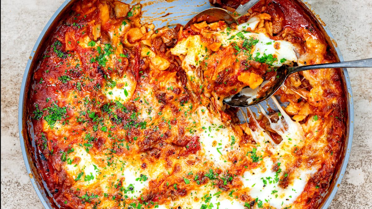 A close-up of a baked skillet lasagna in a round dish, topped with melted cheese, tomato sauce, and sprinkled with chopped parsley. A serving spoon is lifting a portion, revealing pasta layers inside.