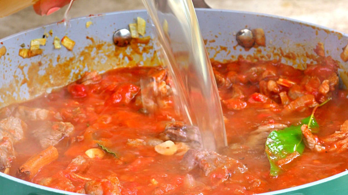 A close-up of a pan filled with ragu sauce, with chopped vegetables, herbs, and bay leaves visible, as a clear liquid is being poured into the mixture.