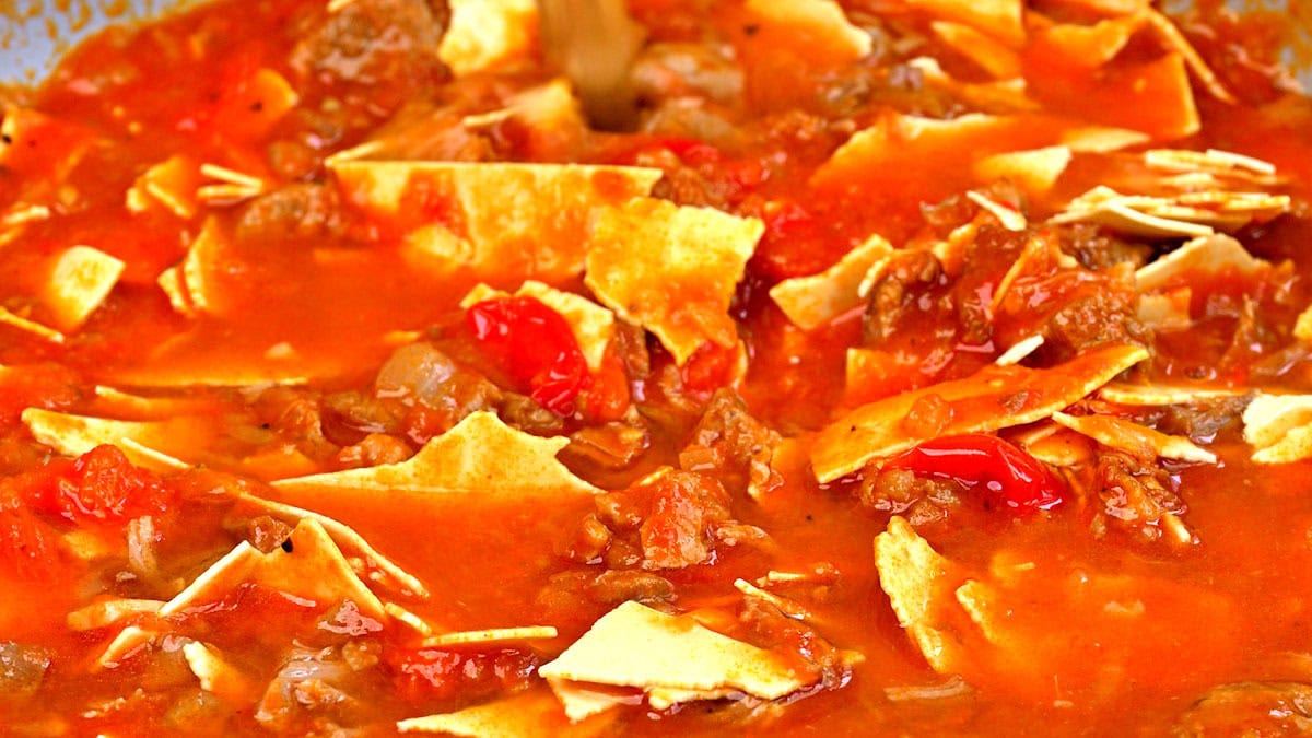A close-up of a hearty ragu with chunks of meat, tomatoes, and pieces of broken up lasagna noodles cooking in a rich, red, tomato-based broth.