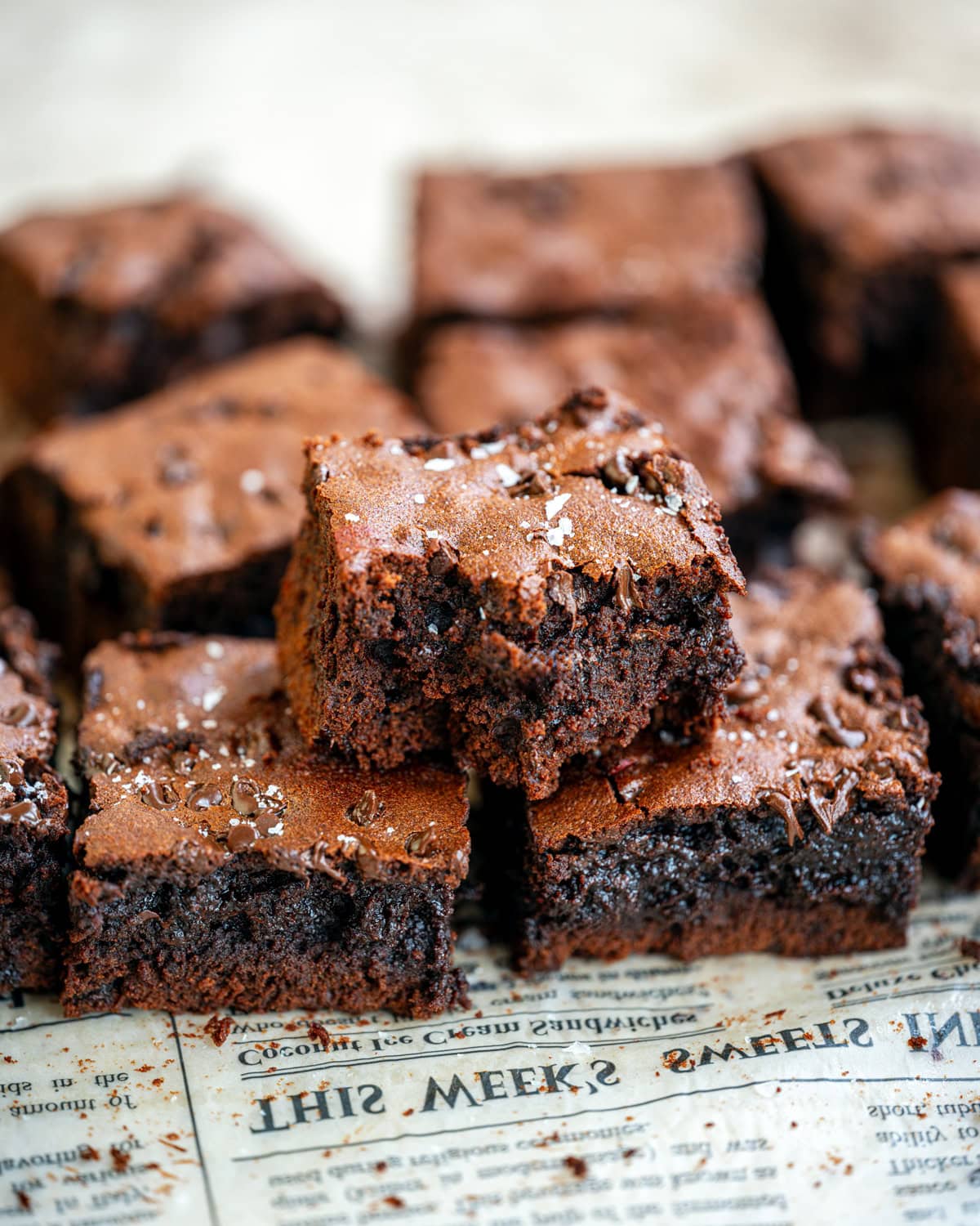 A close-up of several rich, fudgy chocolate brownies stacked on a surface covered with newspaper. One brownie on top has a bite taken out, showing its moist, dense interior.