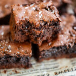 A close-up of chocolate protein brownies stacked on old newspaper, with one brownie showing a bite taken out. The brownies have a rich, fudgy texture and are sprinkled with flaky sea salt.
