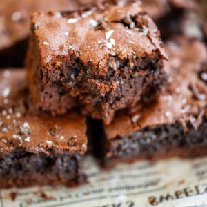 A close-up of chocolate protein brownies stacked on old newspaper, with one brownie showing a bite taken out. The brownies have a rich, fudgy texture and are sprinkled with flaky sea salt.