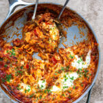 A skillet filled with cheesy lasagne topped with parsley. Two serving utensils rest in the pan, with a portion of the pasta scooped out, revealing a saucy, golden, and slightly browned surface.