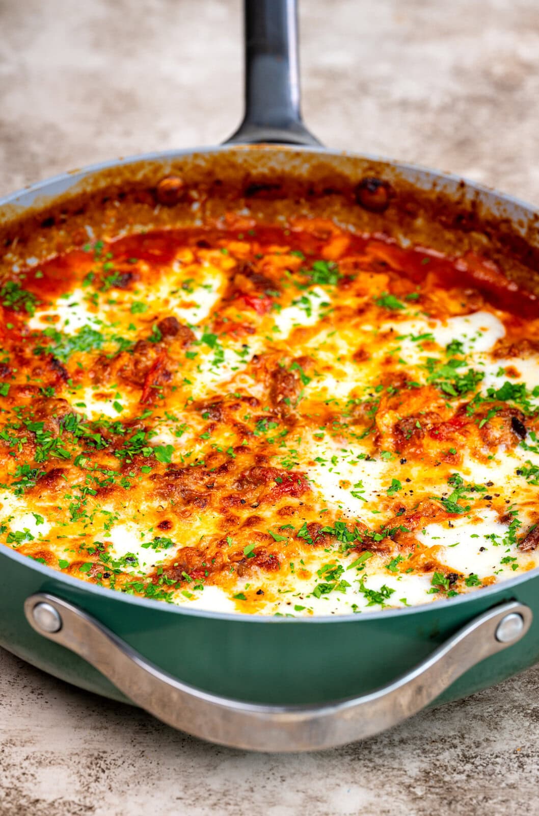 A skillet filled with baked lasagna, topped with melted cheese, browned edges, tomato sauce, and sprinkled with fresh chopped parsley.