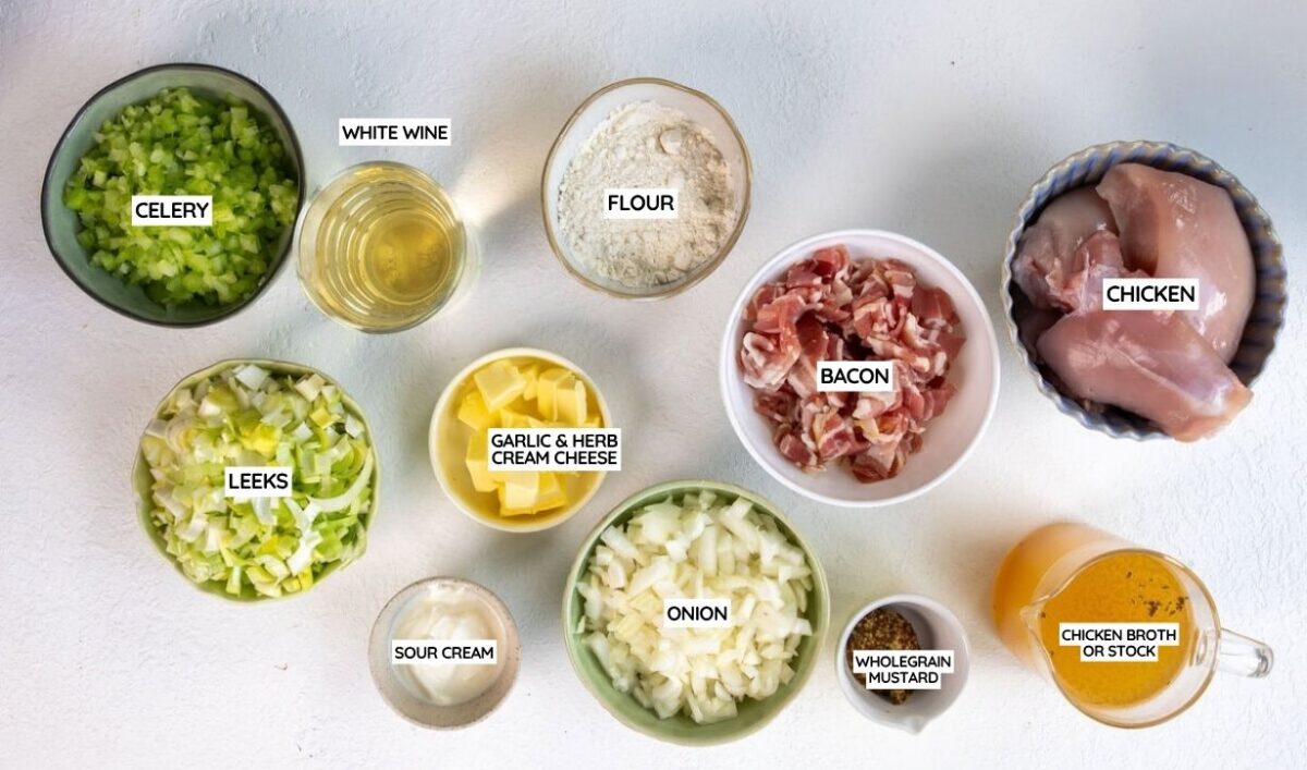 Top-down view of labeled bowls containing celery, white wine, flour, bacon, chicken, leeks, garlic & herb cream cheese, onion, sour cream, wholegrain mustard, and chicken broth or stock on a white surface.