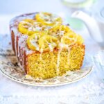 A loaf of lemon poppy seed cake with icing and candied lemon slices on top sits on a decorative plate, with a teapot and teacup blurred in the background.
