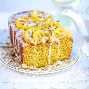 A loaf of lemon poppy seed cake with icing and candied lemon slices on top sits on a decorative plate, with a teapot and teacup blurred in the background.