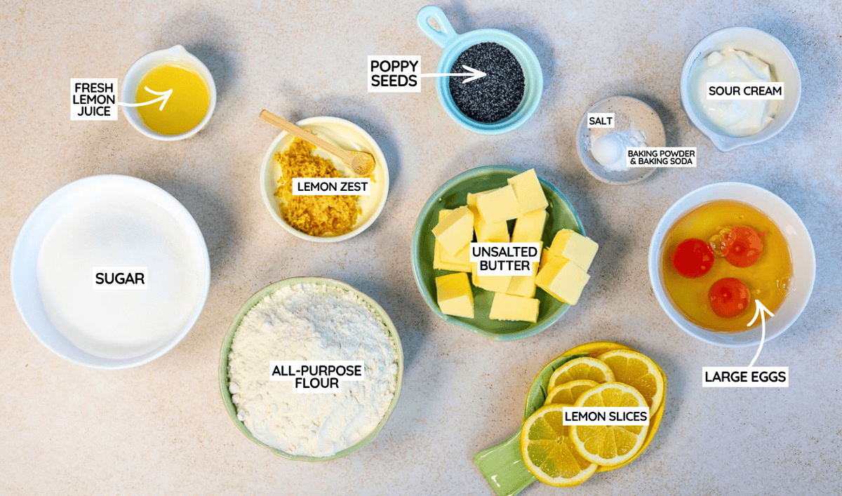 Top-down view of baking ingredients in bowls, including flour, sugar, eggs, butter, sour cream, lemon juice, lemon zest, lemon slices, poppy seeds, salt, and baking powder & baking soda, all labeled for a recipe.