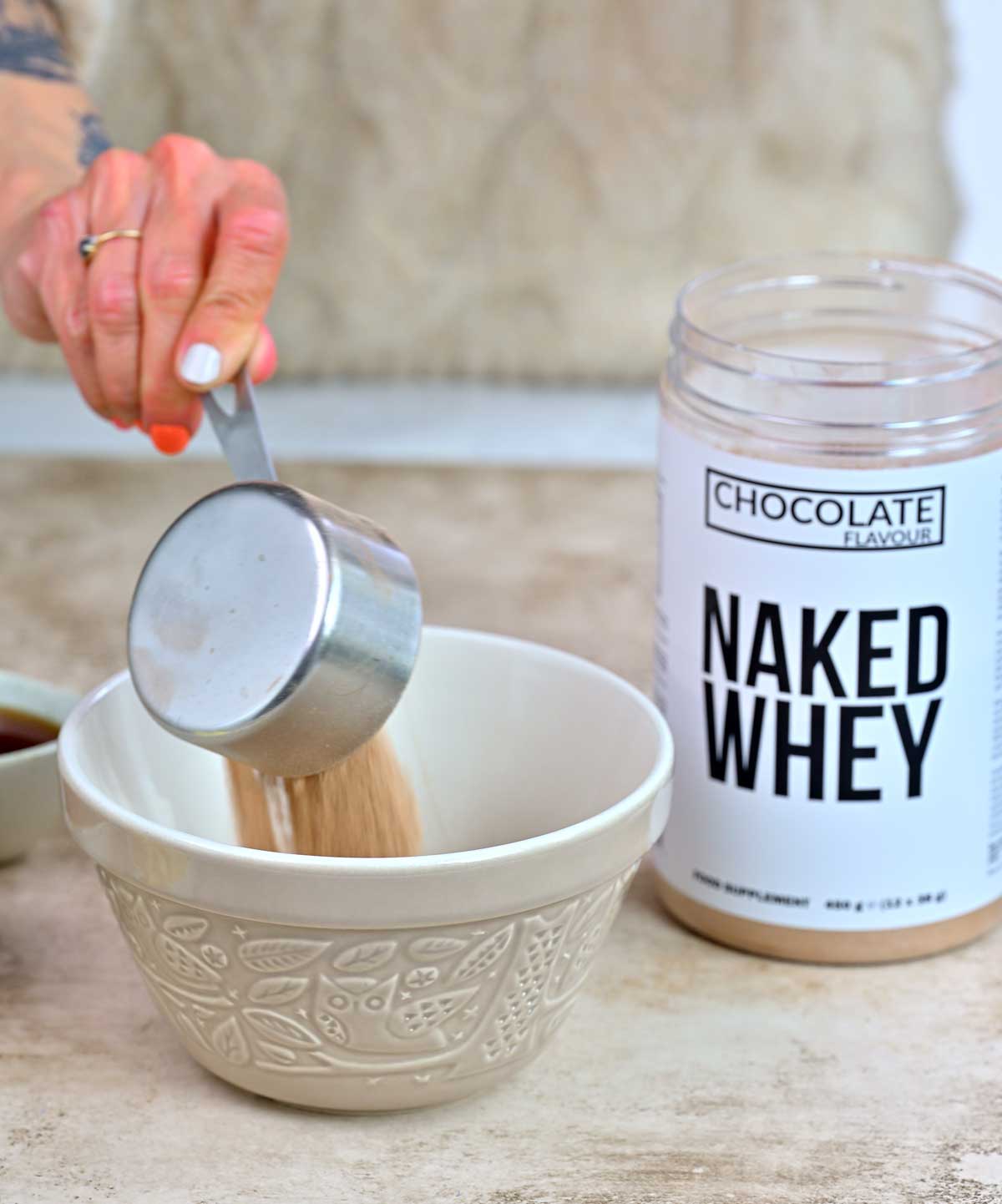 A hand pours a scoop of chocolate-flavored Naked Whey protein powder from a metal measuring cup into a patterned ceramic mixing bowl on a countertop.