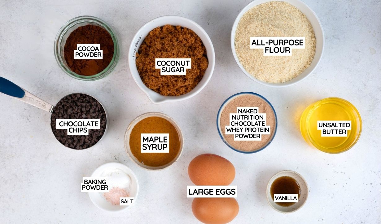 Top-down view of baking ingredients in bowls and cups, labeled: cocoa powder, coconut sugar, all-purpose flour, chocolate chips, maple syrup, protein powder, unsalted butter, baking powder, salt, large eggs, and vanilla.