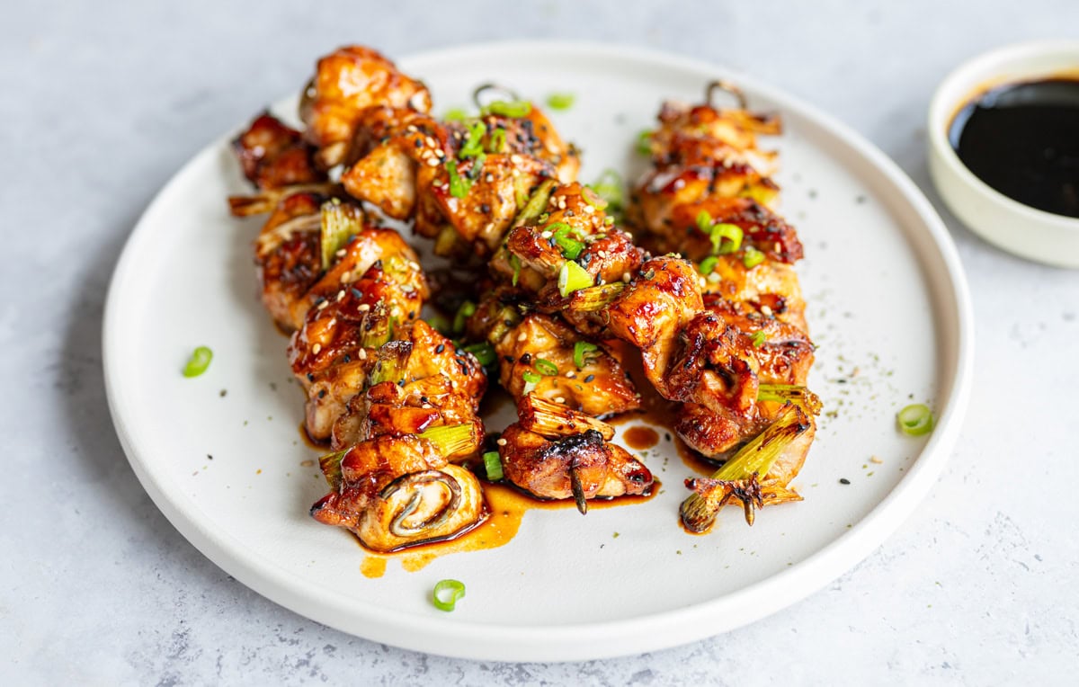 Grilled air fryer chicken skewers (yakitori) glazed with a shiny sauce, garnished with chopped green onions, served on a white plate with a small bowl of dark dipping sauce on the side.
