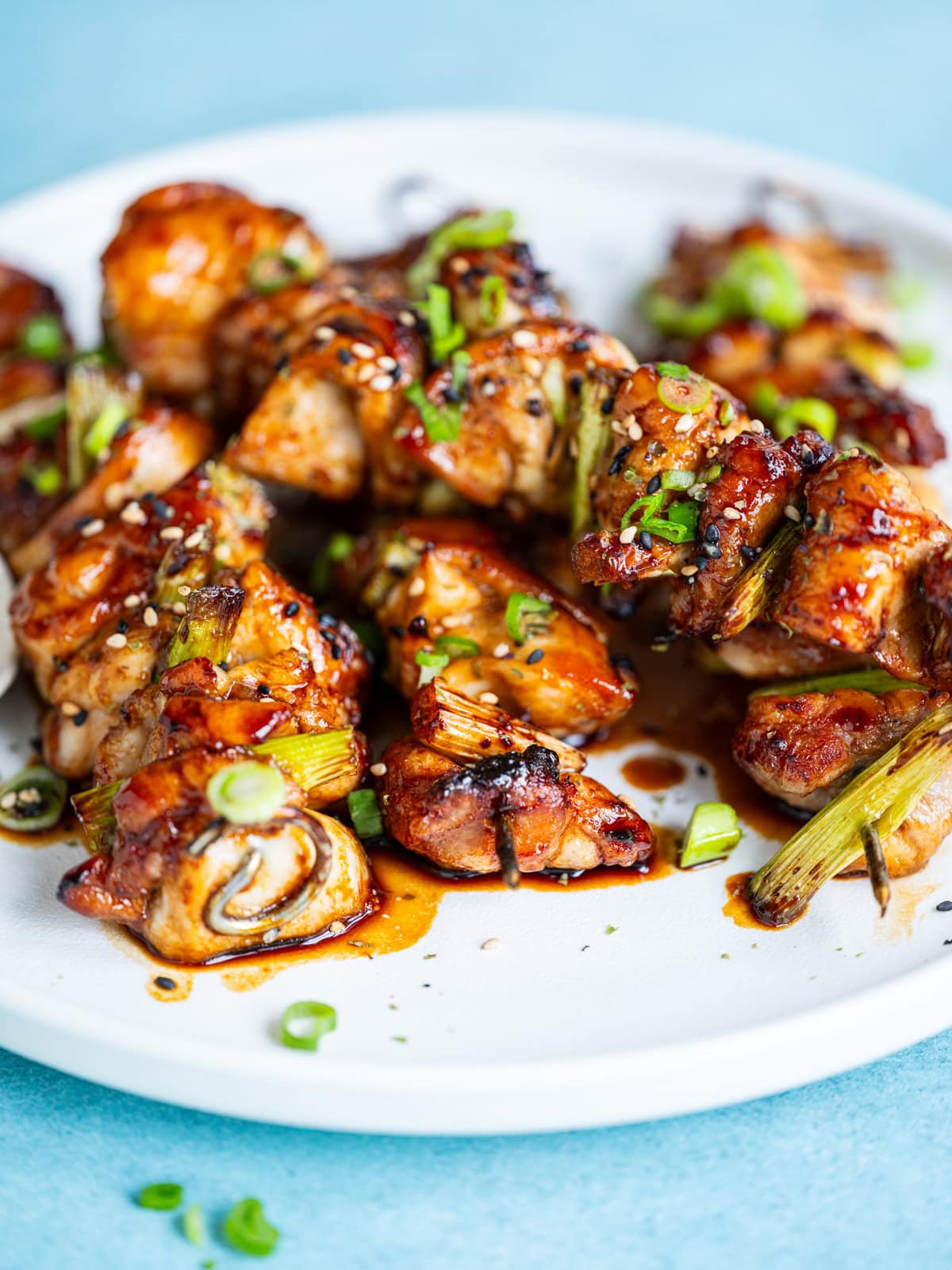 Air Fryer Japanese Yakitori chicken skewers glazed with a shiny sauce, garnished with sliced green onions, served on a white plate against a light blue background.