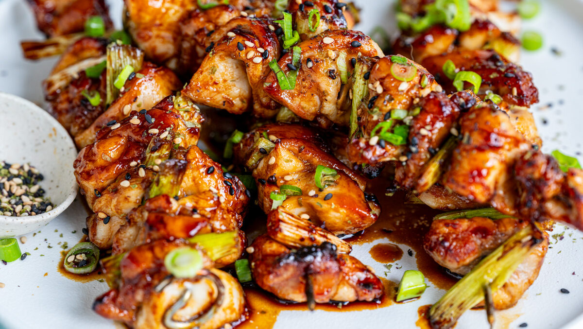 Skewers of grilled chicken glazed with a shiny sauce, garnished with sliced green onions, are served on a white plate with a small dish of seasoning on the side, against a light blue background.