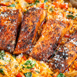 A one pan skillet filled with four Cajun Salmon Orzo fillets atop creamy orzo pasta with spinach and tomatoes, all topped with grated cheese.