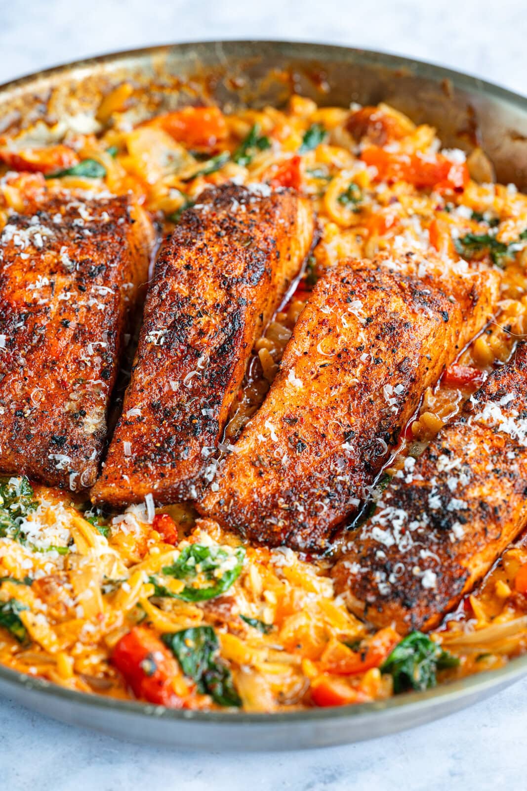 A one pan skillet filled with four Cajun Salmon Orzo fillets atop creamy orzo pasta with spinach and tomatoes, all topped with grated cheese.