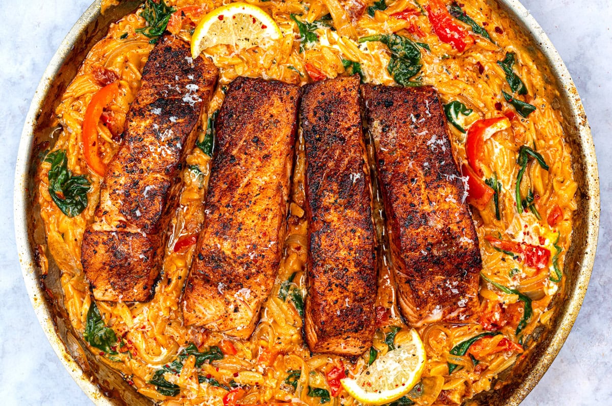 Four blackened cajun salmon fillets on top of creamy pasta with spinach, red bell peppers, and lemon slices in a skillet. The dish is garnished with herbs and grated cheese.
