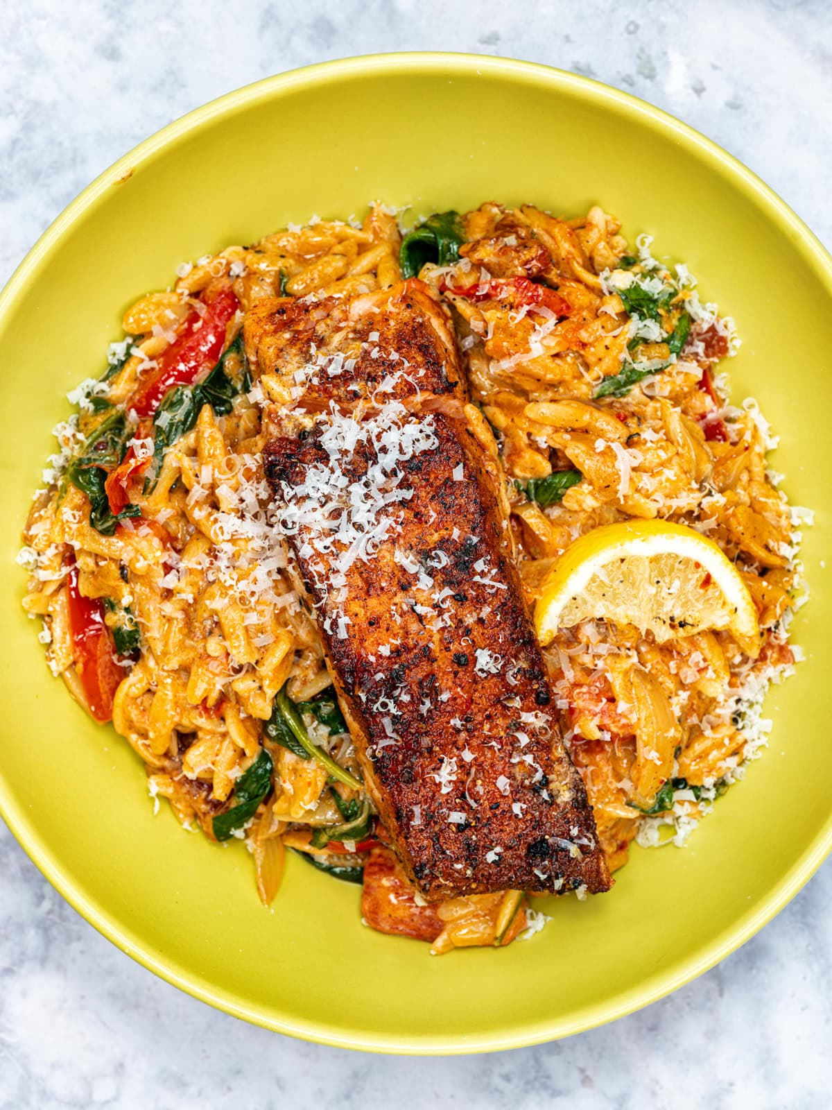 A yellow plate with creamy orzo, spinach, and red bell peppers, topped with a blackened salmon fillet, grated cheese, and a slice of lemon.