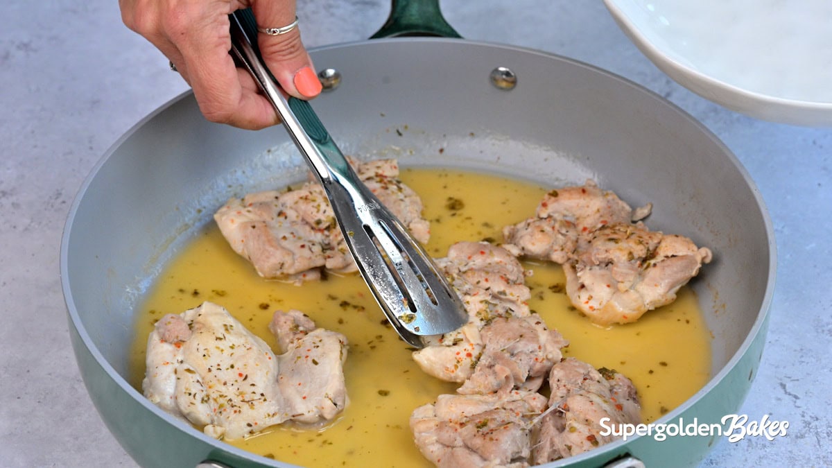 A hand uses tongs to turn seasoned chicken thighs cooking in a pan with yellow sauce. The pan is on a gray surface, and Supergolden Bakes is written in the corner.