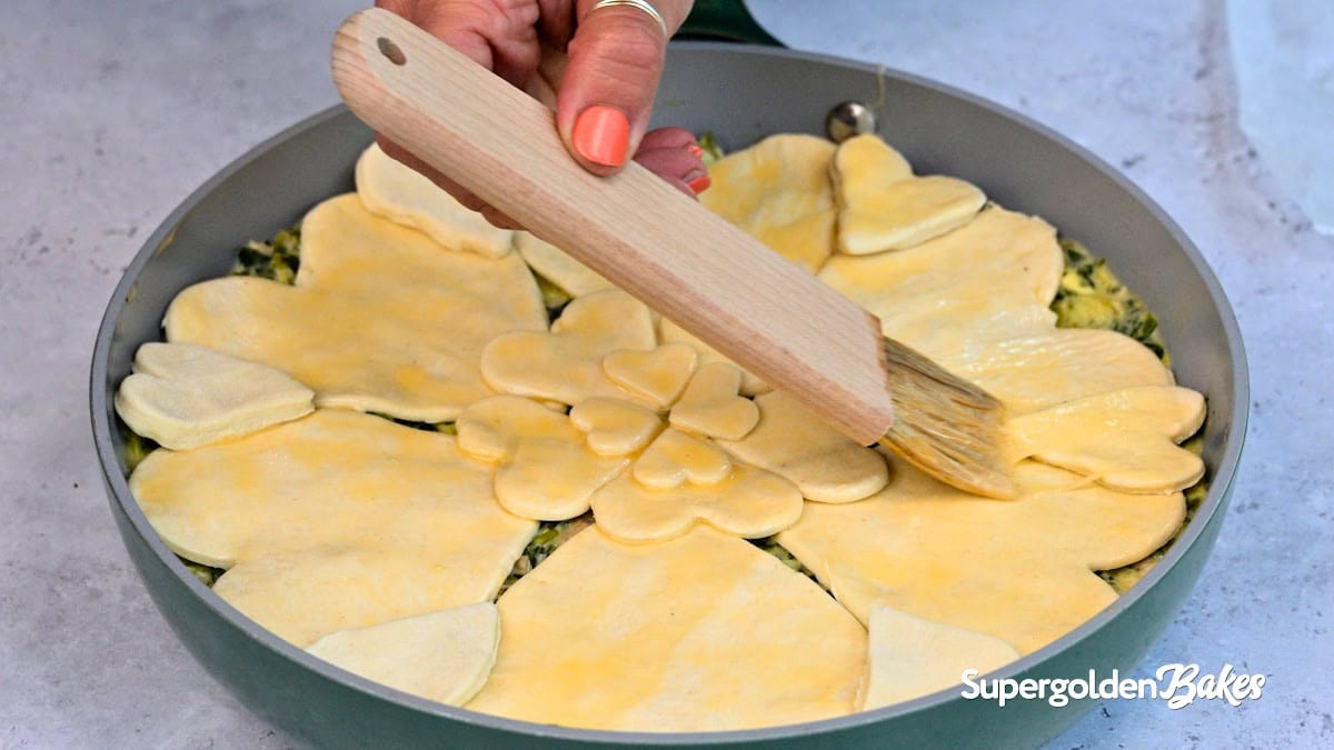 A hand uses a brush to apply egg wash to a pie topped with heart- and flower-shaped pastry cutouts in a round pan. The logo Supergolden Bakes appears in the bottom right corner.