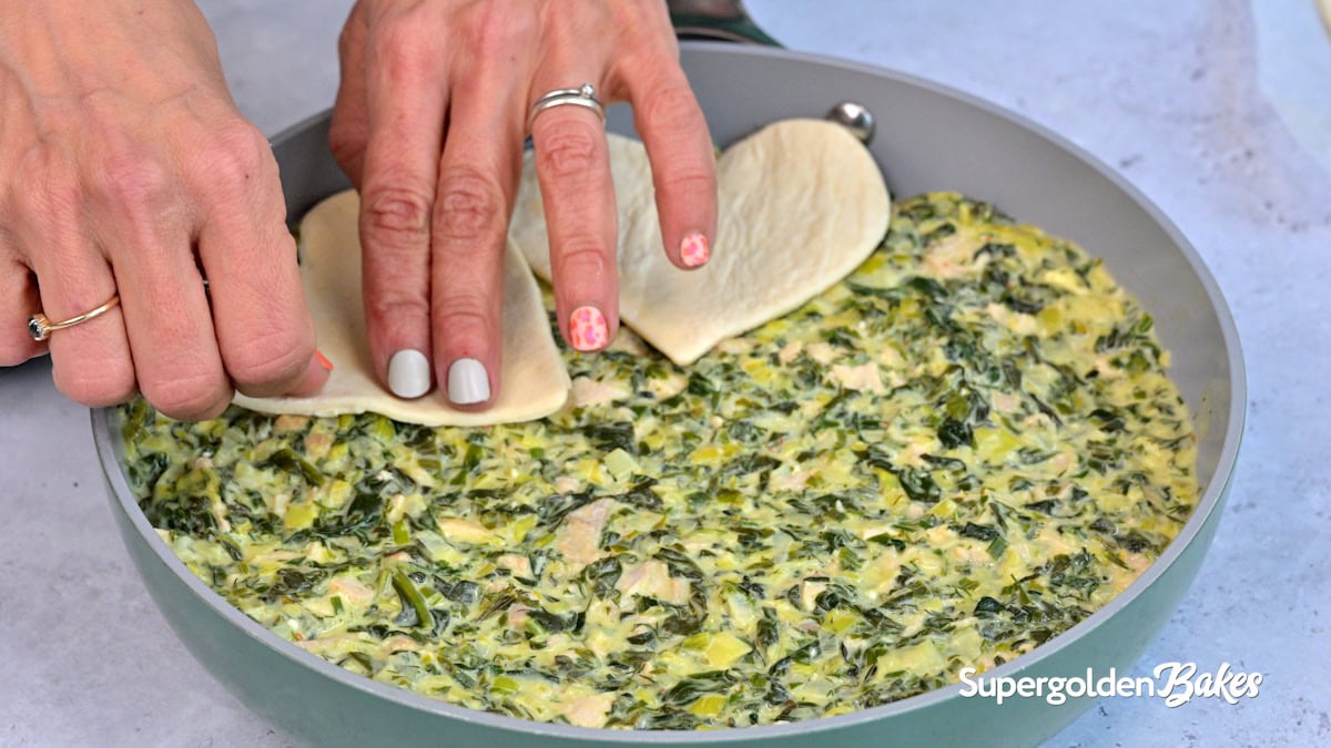 Close-up of hands placing pieces of puff pastry over a creamy spinach and chicken mixture in a round pan. The pan is labeled “Supergolden Bakes.” The person has decorated nails and wears rings.