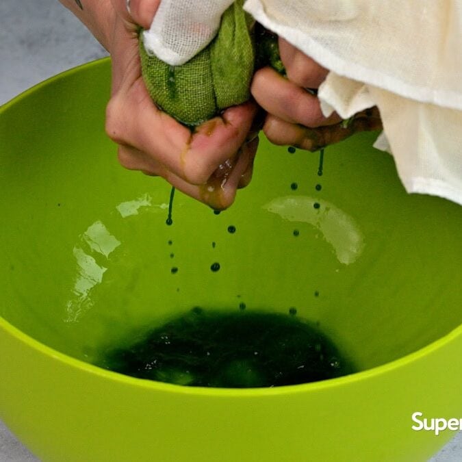 A person squeezes spinach in cheese cloth over a bright green bowl, straining green liquid through a white cheesecloth. The bowl sits on a gray surface, with Supergolden Bakes written in the corner.