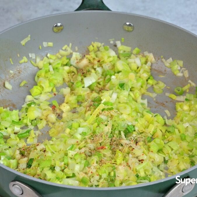 Chopped leeks and seasonings are sautéing in a large, light gray frying pan on a stovetop. The pan’s handle and part of a brown cutting board are visible. Supergolden Bakes is written in the corner.