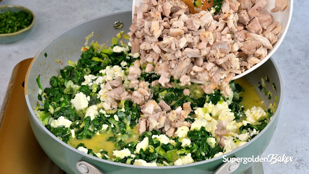 A person adds chopped cooked chicken from a white bowl into a pan filled with chopped spinach, scrambled eggs, and feta cheese. The pan is on a stove, and the scene is from Supergolden Bakes.