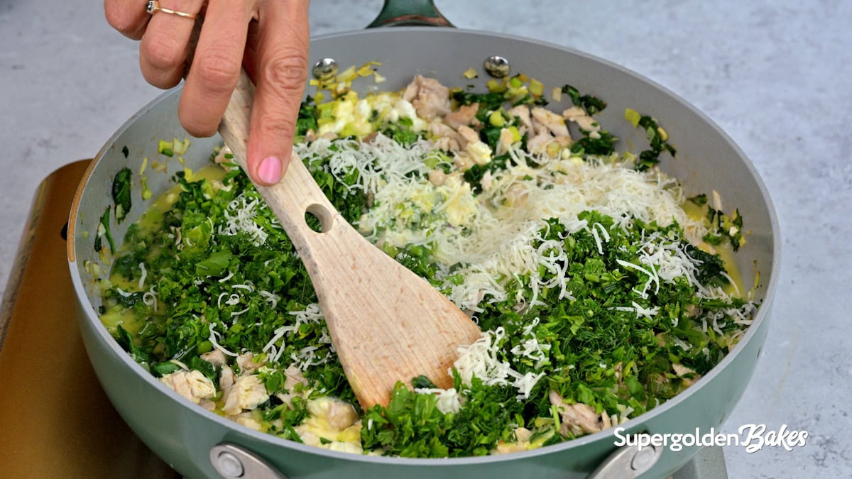 A hand stirs a mixture of chopped greens, shredded cheese, and chicken in a large skillet with a wooden spoon. The scene is viewed from above, and the pan sits on a gray countertop. Supergolden Bakes is written in the corner.