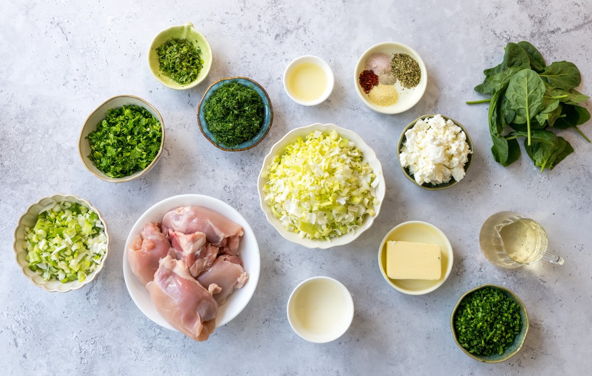 Various ingredients for a chicken and spinach pot pie recipe are arranged on a light surface, including raw chicken, chopped greens, cabbage, spinach, herbs, spices, cottage cheese, butter, oil, and a bowl of liquid, possibly broth or wine.