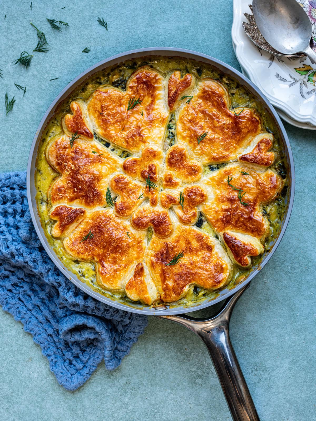 A skillet filled with a golden-brown, flower-shaped Spanakopita pot pie with chicken topping a creamy vegetable filling, garnished with fresh herbs, sits on a blue textured cloth next to stacked plates and silver serving utensils.