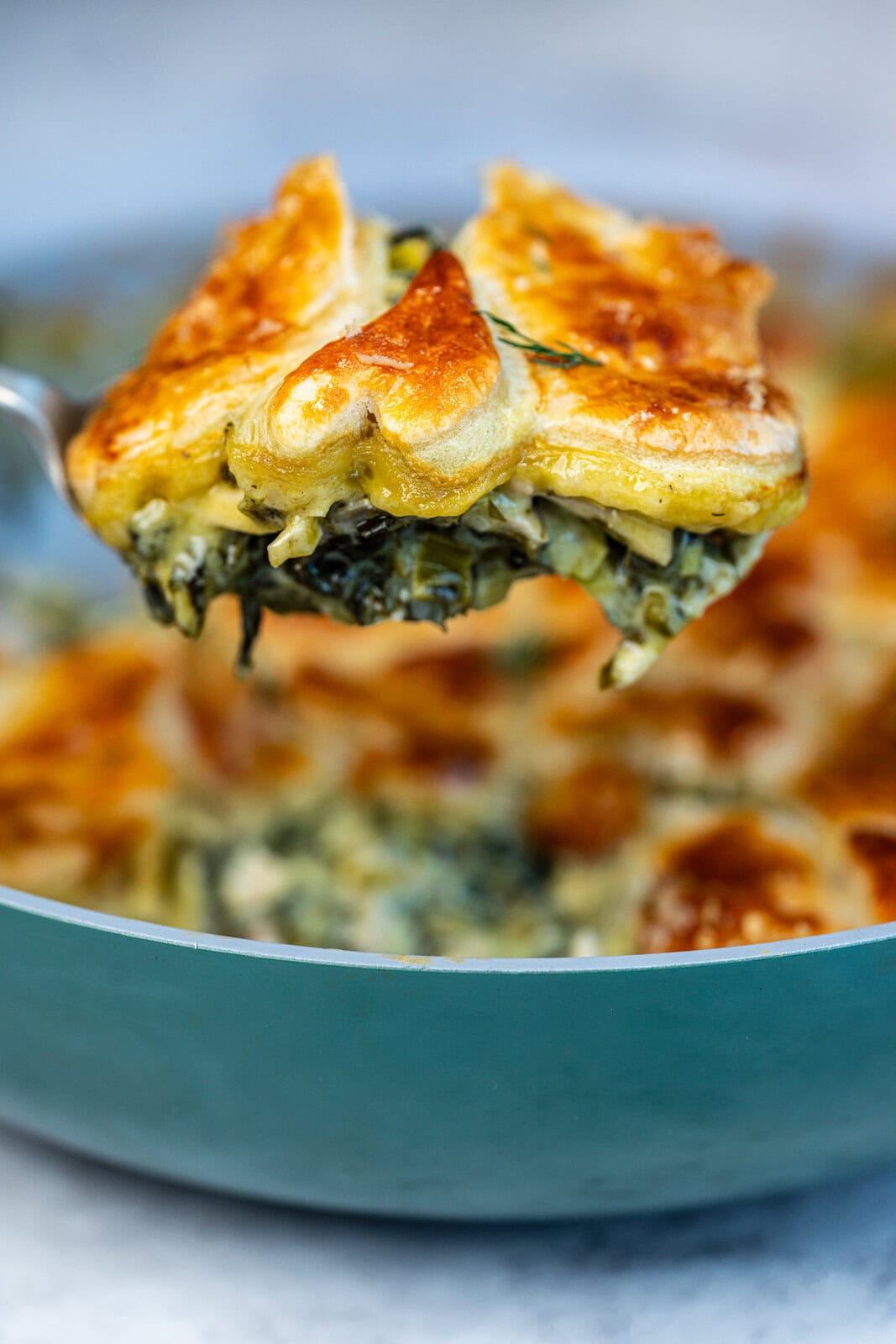 A close-up of a spoon lifting a serving of creamy spinach pie topped with a golden, flaky puff pastry shaped like a heart from a teal baking dish.