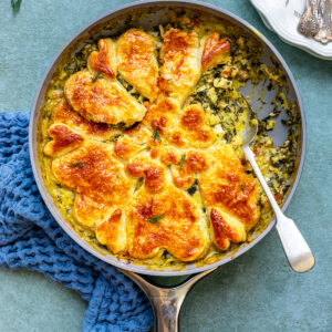 A skillet filled with a golden-brown, puff pastry-topped Spanakopita pot pie with chicken, with a serving spoon and blue textured cloth underneath. A plate with two forks sits beside the skillet on a green surface.