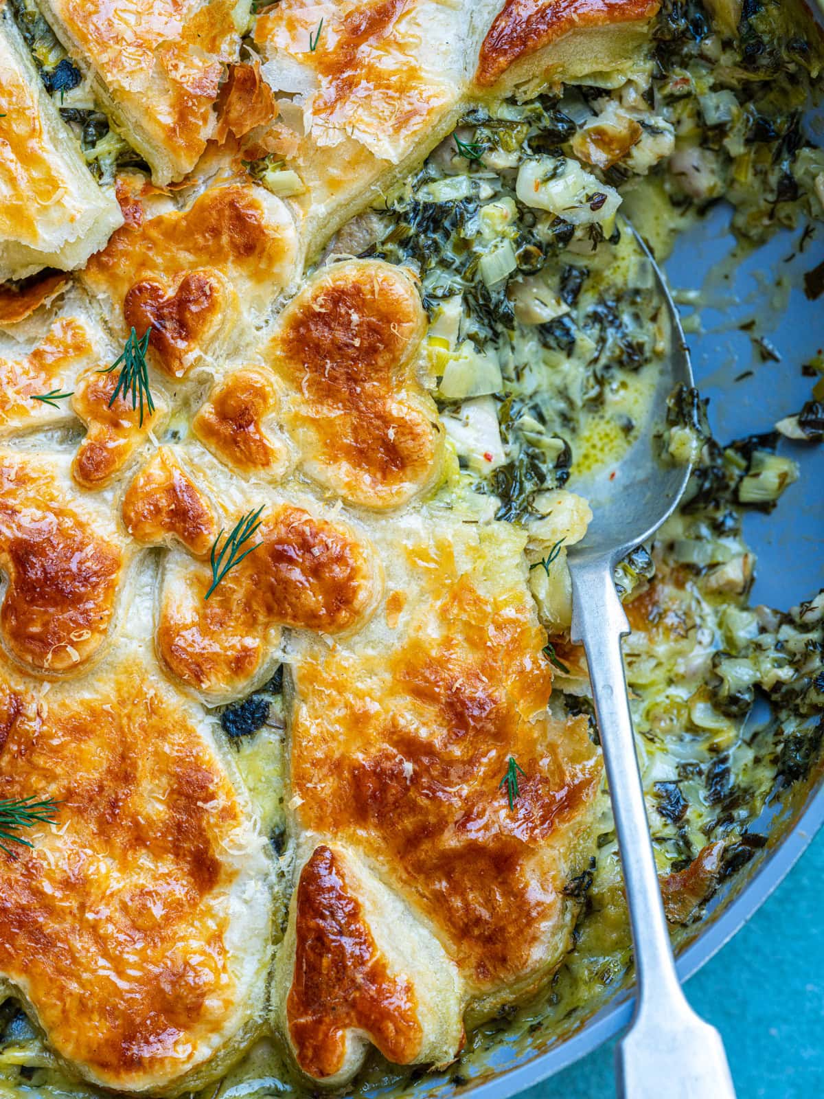 Golden, flaky puff pastry tops a creamy, vegetable-filled pie with herbs, shown in a baking dish. A silver spoon rests inside, revealing the savory filling under the browned pastry crust.